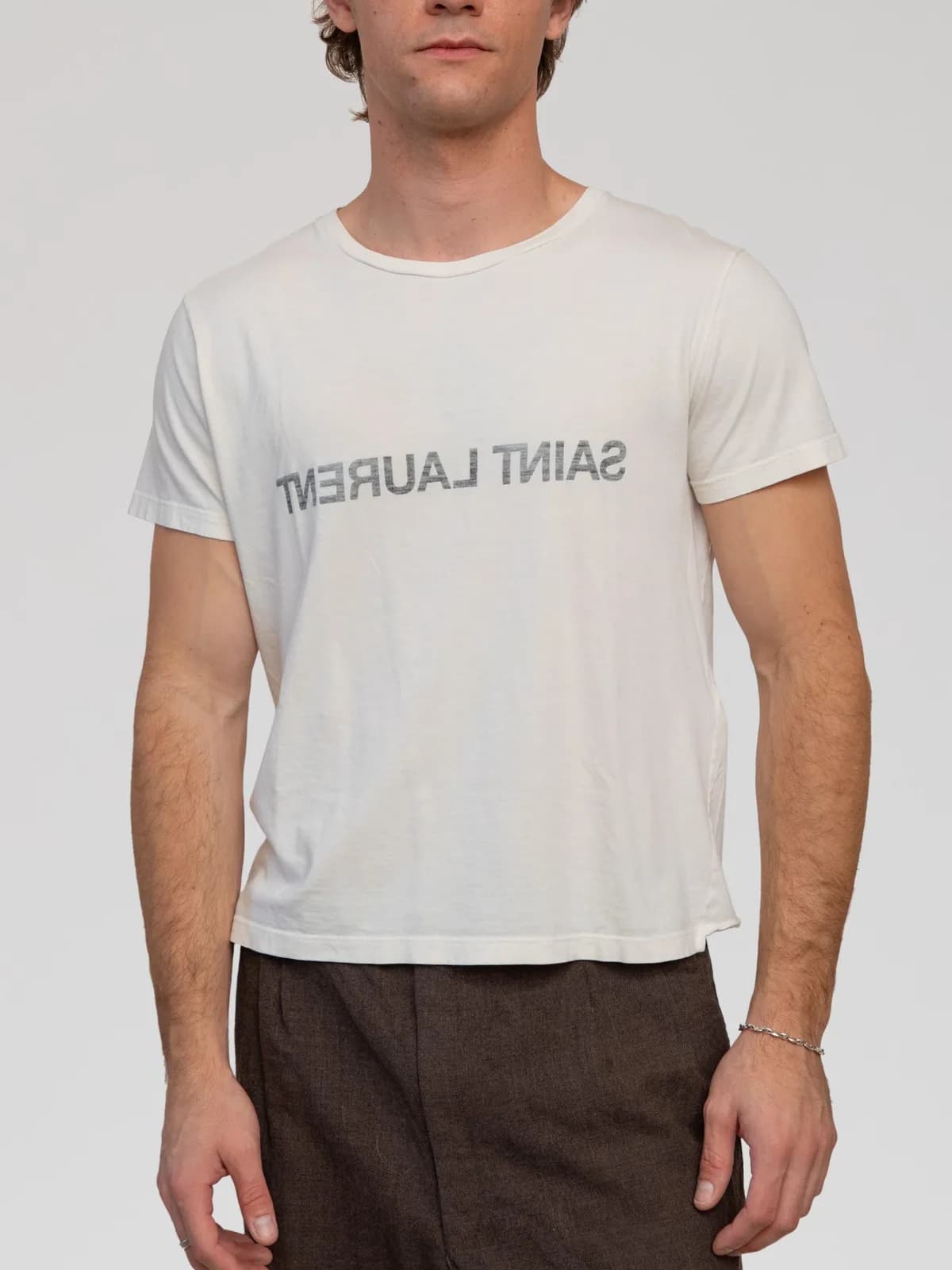 Anthony Vaccarello Era Saint Laurent Paris off-white short-sleeve jersey t-shirt crafted in lightweight cotton, featuring a distinctive reverse "SAINT LAURENT" logo printed in mirrored dark grey lettering across both the chest and back, finished with a classic crew neckline and a raw-edge back hemline. - Image 1