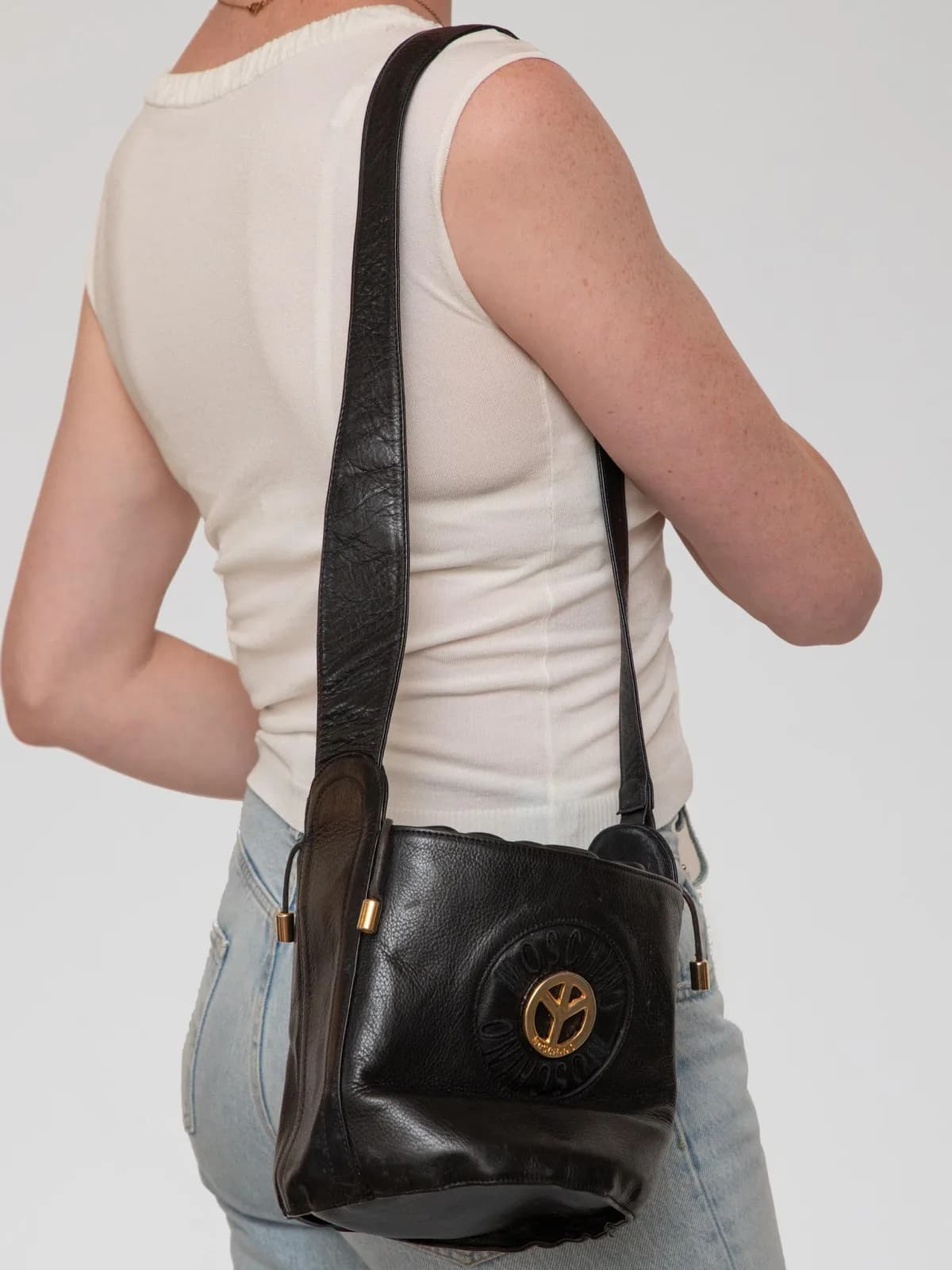 Crafted in supple black grained leather, this vintage Moschino drawstring bucket bag features a bold gold-tone peace sign hardware emblem surrounded by an embossed logo ring, drawstring side ties, and a comfortable flat shoulder strap for a playfully chic archival style. - Image 1