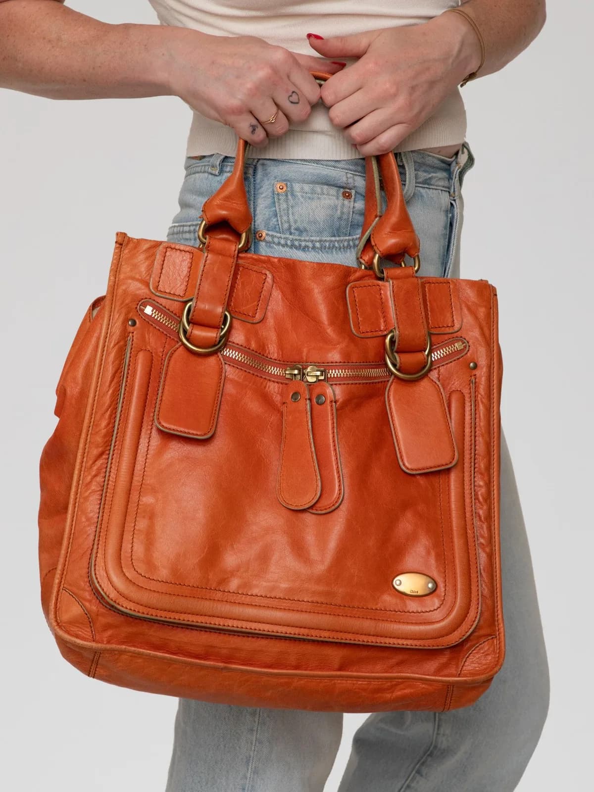 Crafted in a vibrant burnt orange leather with aged brass hardware, this iconic vintage Chloé Bay bag features distinctive front zip pockets with elongated leather pulls, sturdy rolled handles, and an oval logo plaque for a relaxed, Y2K-era bohemian luxe feel. - Image 1