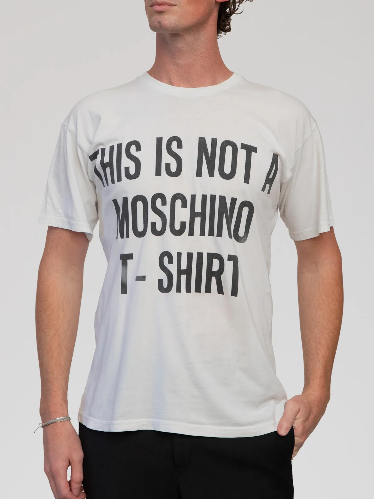 Jeremy Scott Era Moschino Couture white cotton short-sleeve t-shirt featuring the iconic surrealist slogan "THIS IS NOT A MOSCHINO T-SHIRT" printed in large black block lettering across the front. Designed with a relaxed fit and a ribbed crew neck collar. Made in Italy. - Image 1