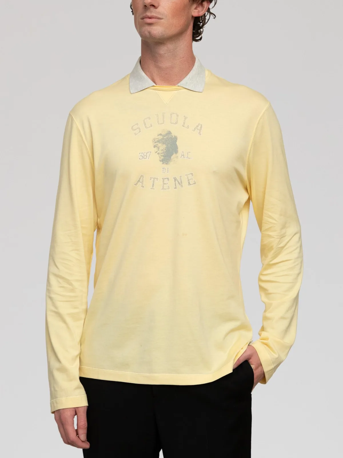 Brunello Cucinelli long-sleeve polo shirt crafted in pale light yellow fine cotton jersey. Features a light heather grey contrasting flat-knit polo collar with a V-insert insert detail, long sleeves, and a straight hem. Accented on the chest with a faded graphic print reading "SCUOLA DI ATENE 387 A.C." surrounding a classic bust graphic. Made in Italy. - Image 1