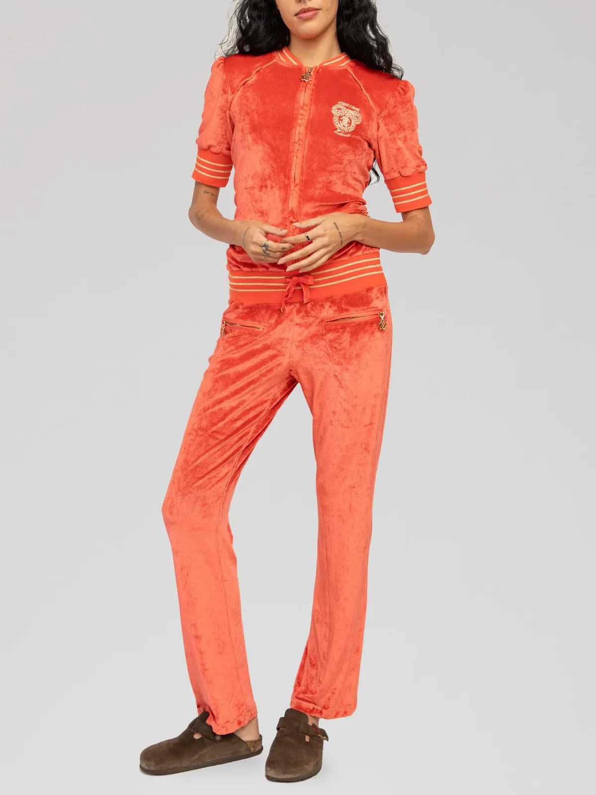 Roberto Cavalli two-piece crushed velour tracksuit set in coral orange featuring a short-sleeve zip-up jacket with gold striped ribbed cuffs, collar, and hem, an embroidered crest logo at the chest, front zipped pockets with branded charms, and matching drawstring straight-leg track pants. - Image 1