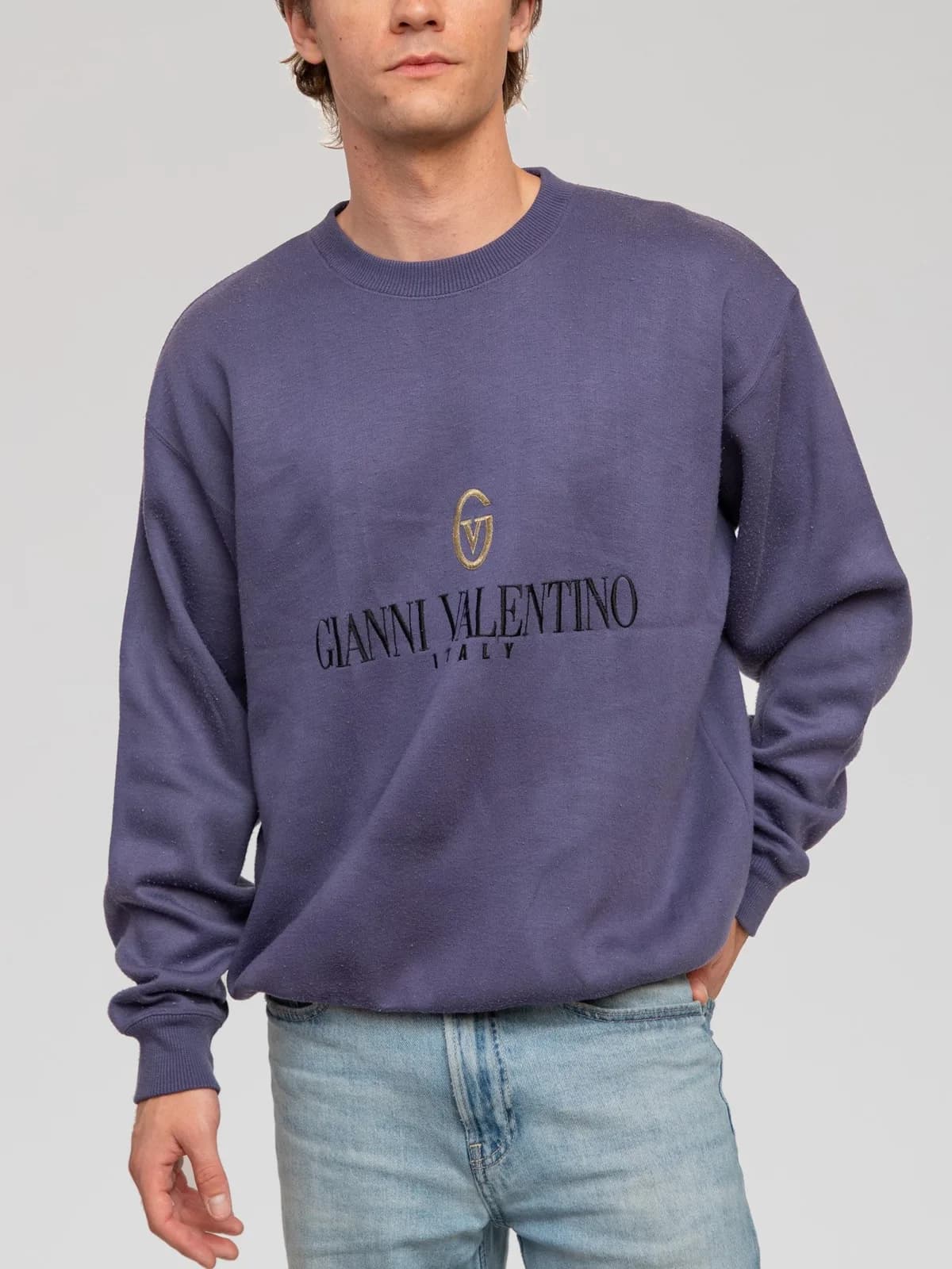 Vintage Gianni Valentino long-sleeve fleece sweatshirt in a muted slate blue hue. Features an embroidered gold "GV" logo emblem and black "GIANNI VALENTINO ITALY" branding across the chest. Designed with a relaxed, slouchy silhouette, dropped shoulders, a ribbed crewneck collar, and ribbed cuffs and hem. - Image 1